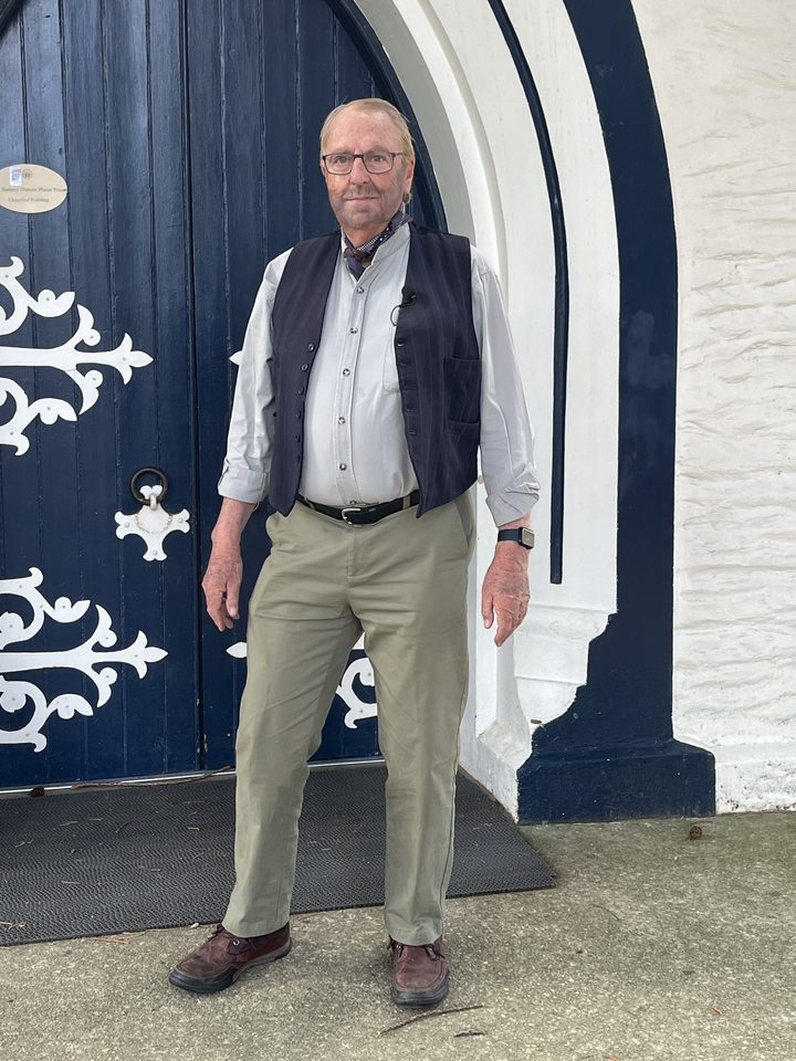 Peter Doyle as William Henry Homer, in a dark waistcoat and cravat, standing at a blue arched door with white ironwork