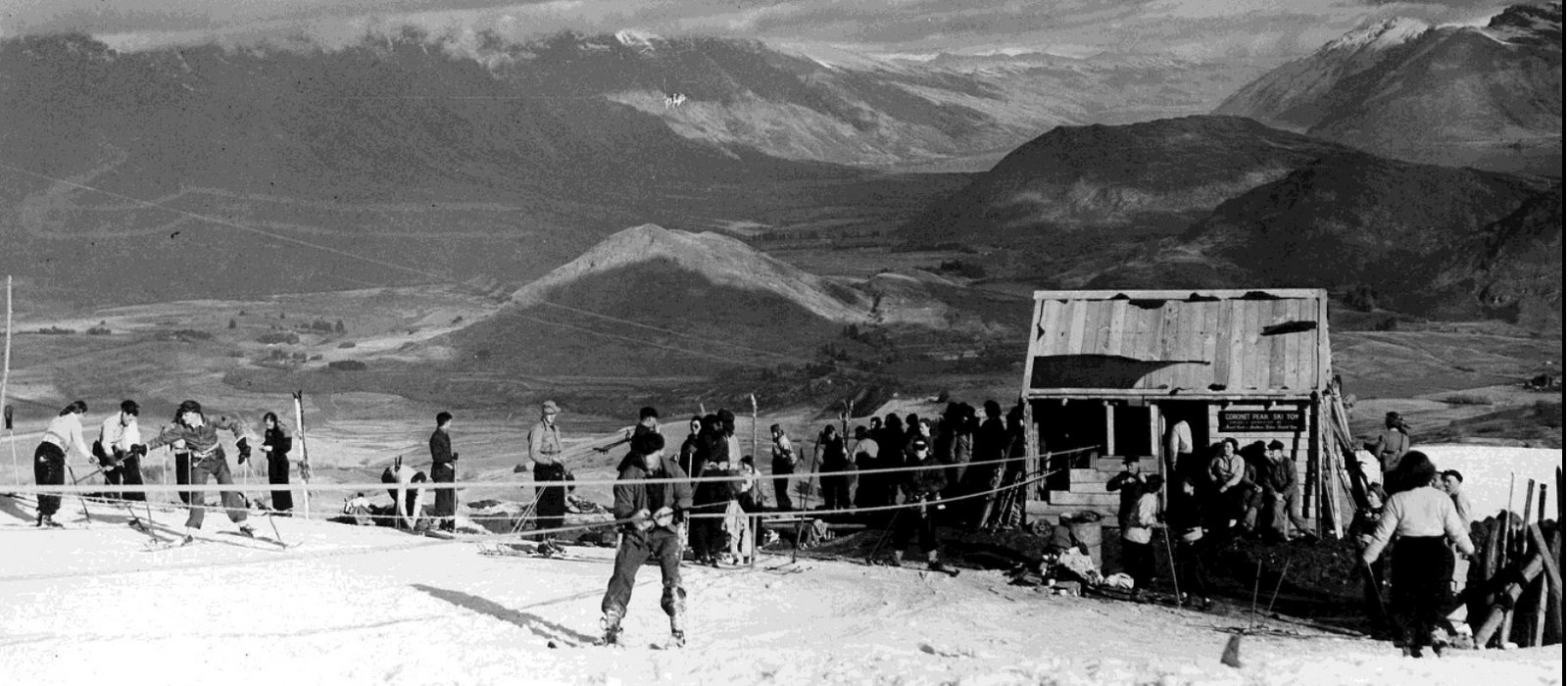 Photograph of skiers in woollen clothing lined up along a rope tow on an open snow slope, with a rough boarded tow shed and a crowd of skis and figures at the right and the dark ranges and valleys of the Whakatipu basin below