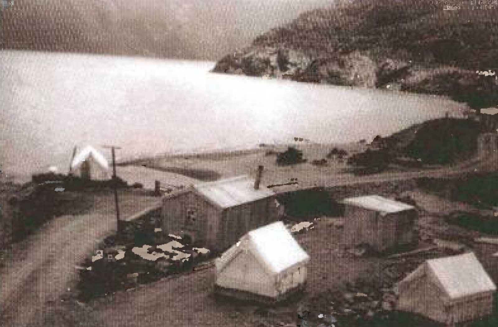 Photograph looking down on a road-camp of about six huts and tents on a bench above a long lake, with a dirt road curving through the camp and a bush-covered spur running out into the water