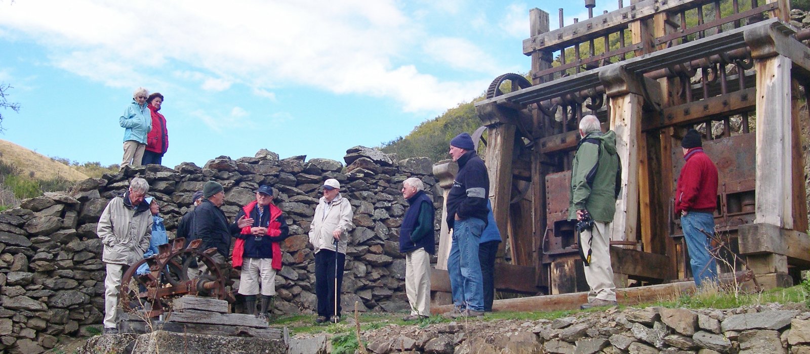 Colour photograph of a dozen Society members in jackets and hats gathered before the timber frame and rusted wheels of a restored stamper battery against a dry-stone wall, with hills and cloud behind