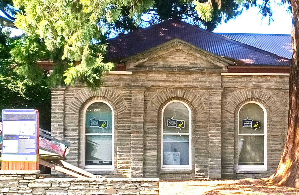 Colour photograph of the stone library building with three arched windows under a pediment and a sequoia, Citizens Advice Bureau signs in the windows and an information panel at the left
