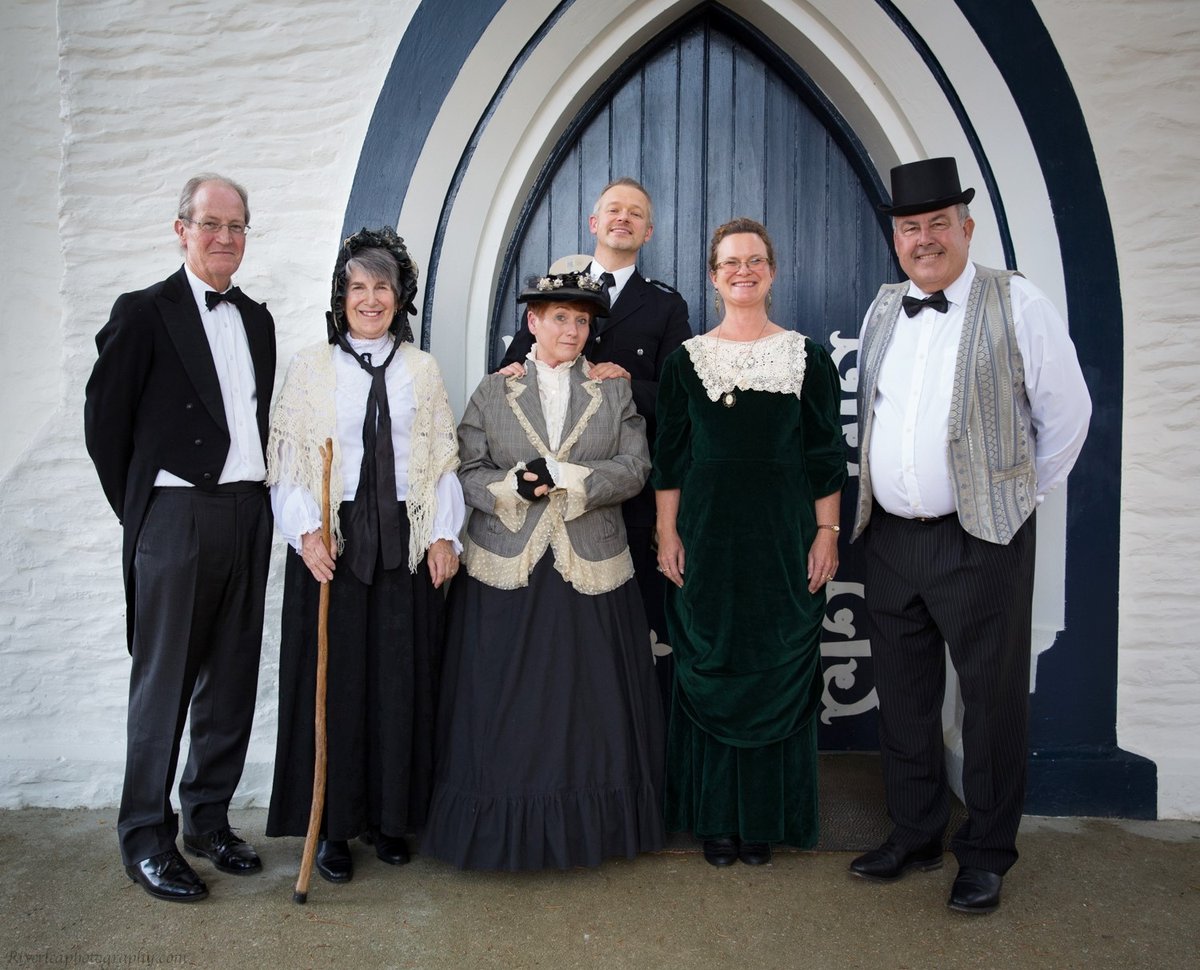 Six people in Victorian costume, among them a policeman, a woman in a bonnet with a walking stick and a man in a top hat, posed in front of a pointed church door