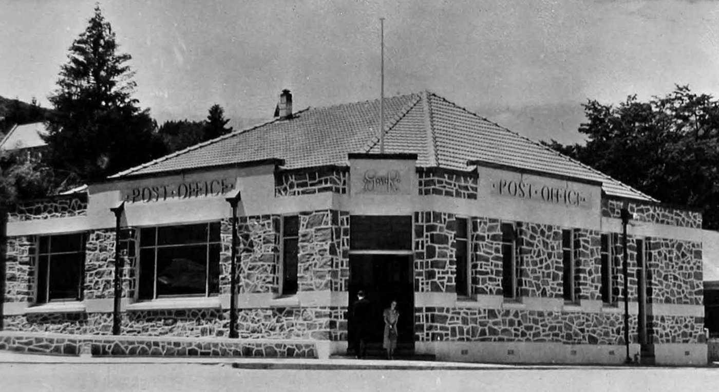 Photograph of a single-storey building on a street corner, its walls faced in rough-cut stone with pale mortar, a deep tiled hipped roof, Post Office lettered on the parapets, a recessed doorway with two people at it, and trees behind