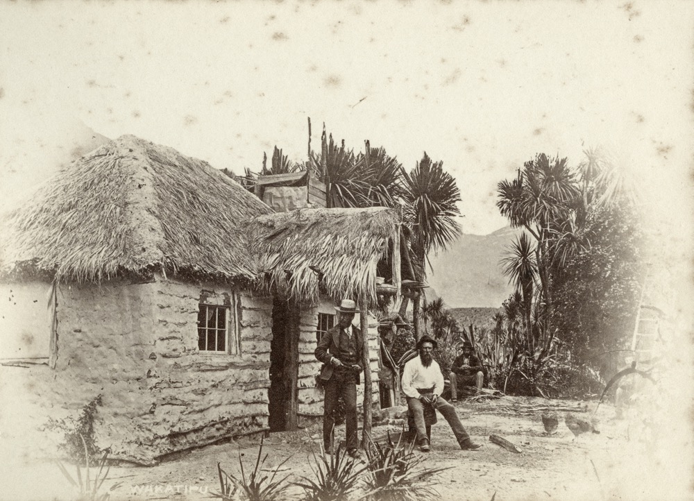 Early photograph of a single-storey cottage with earthen walls and a deep thatched roof, cabbage trees and scrub behind it and three men at the doorway