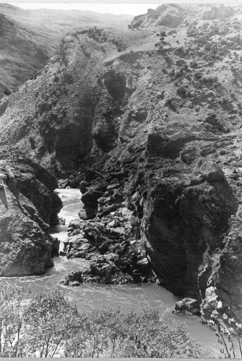Photograph looking down into a rocky gorge where the Kawarau River narrows to a churning channel between high schist bluffs, with tussock hills above and shrubs in the foreground