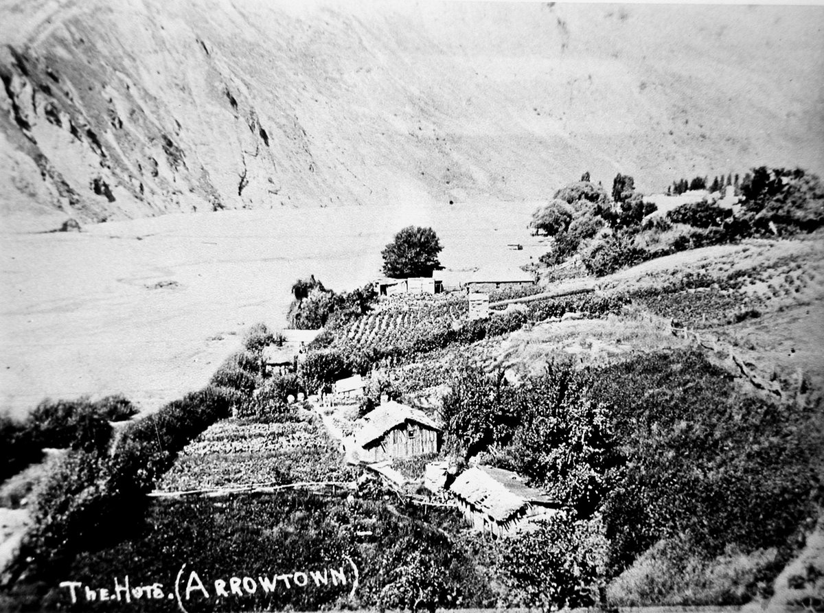 Early photograph looking down over terraced garden plots and thatched huts on a bush-covered slope above a river flat, steep hills behind, captioned in white lettering 
