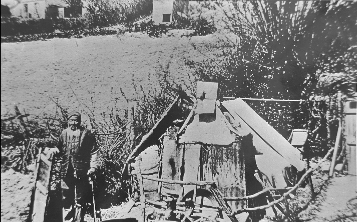 Early photograph of a Chinese man in a cap and long coat standing with a stick beside a low hut of boards and sacking, bare trees and a garden plot behind