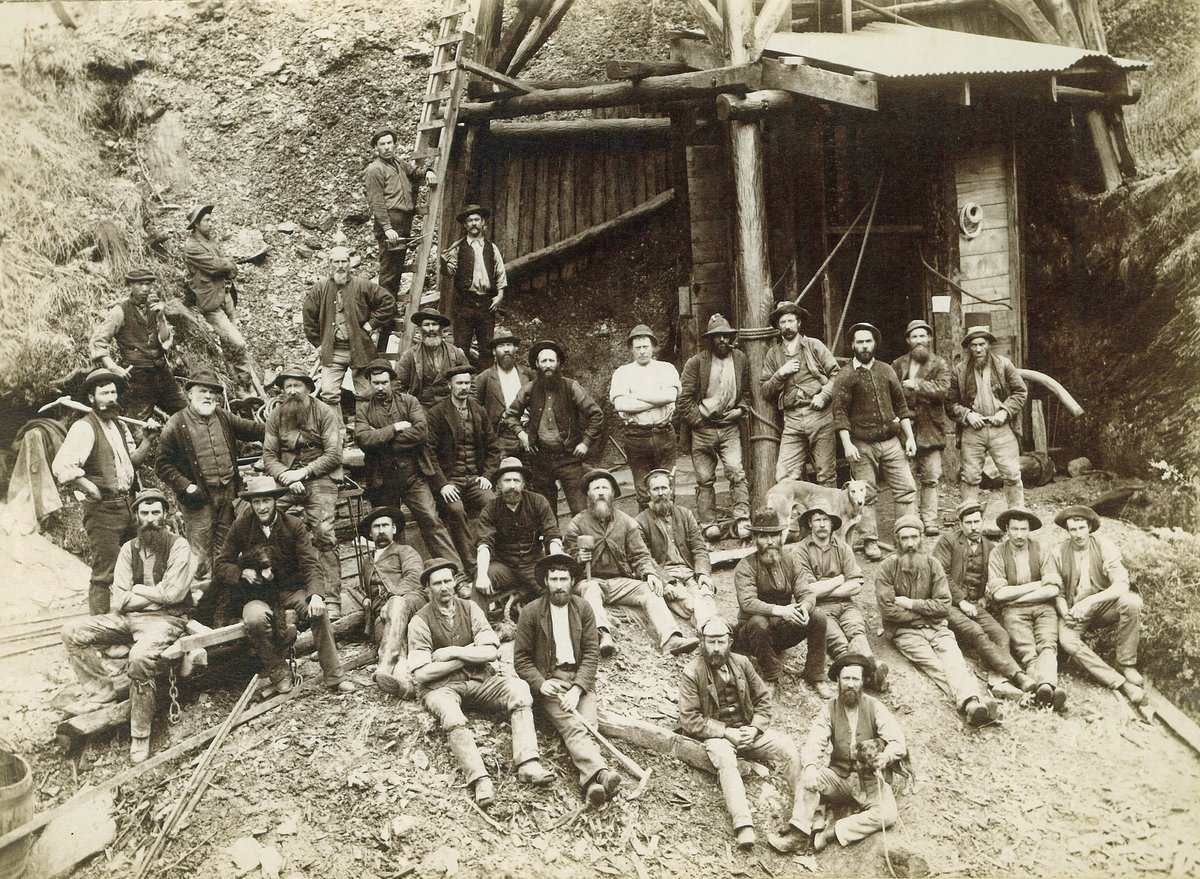 Sepia photograph of about forty bearded miners in hats and waistcoats, standing and sitting on the ground in front of a timber mine entrance and headframe, two dogs among them