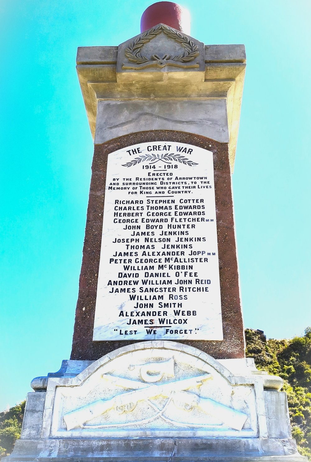 Stone war memorial photographed from below against a blue sky, its marble tablet headed The Great War 1914 to 1918 and listing eighteen names, with crossed rifles carved on the base