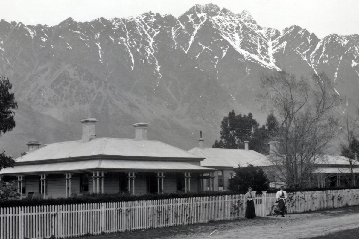 Photograph of a single-storey hospital building with a verandah and picket fence, two people at the gate with a bicycle, and the snow-streaked Remarkables filling the sky behind