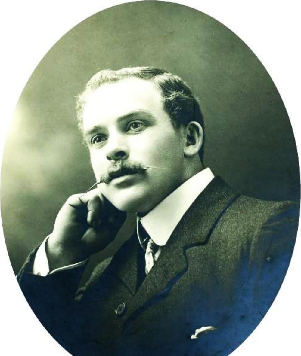 Studio portrait in an oval frame of a young man with fair wavy hair and a moustache, in a dark suit and high collar, his chin resting on his hand