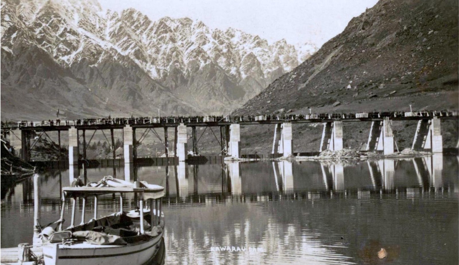 Photograph across still water to a long low bridge on concrete piers, its deck lined with people, with a bare scree hillside on the right, the snow-streaked Remarkables behind and a moored launch in the foreground