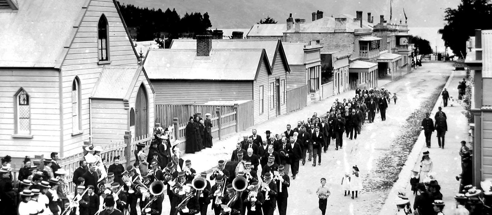 Photograph from above of a brass band leading a procession of men down a dirt street past a weatherboard church with a bell tower, crowds on both footpaths, and shops and hotels running down to the lake and mountains