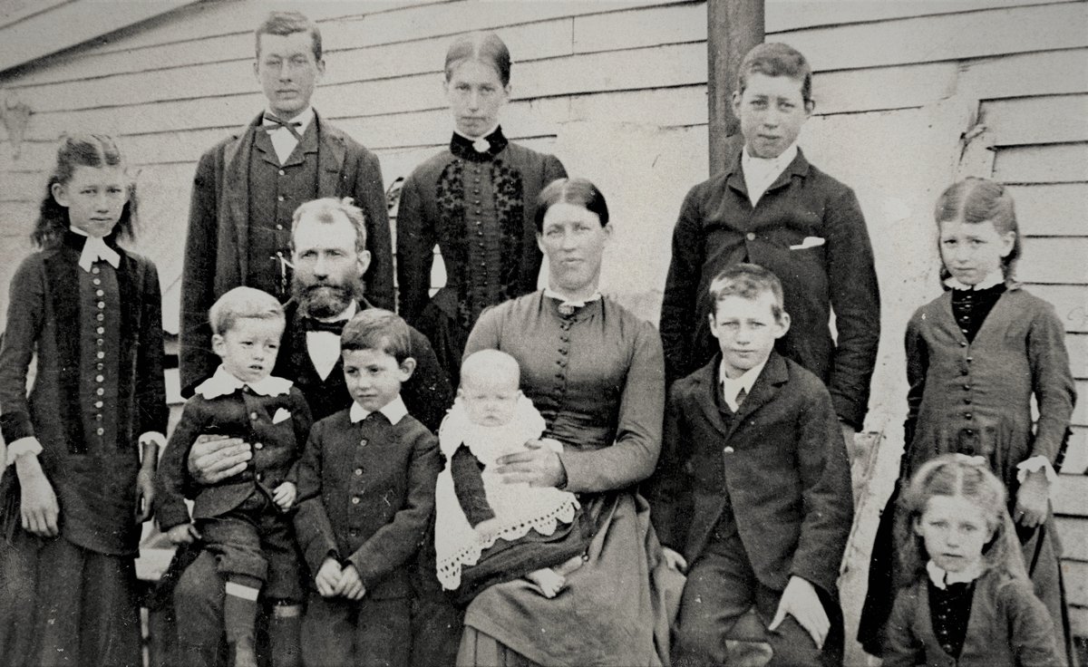 Family portrait against a weatherboard wall: a bearded father and a mother holding a baby, with ten children from a young man in a bow tie to small girls in dark buttoned dresses