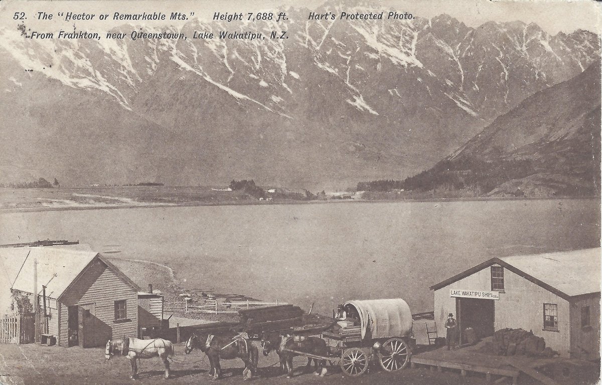 Postcard view across Lake Whakatipu to the snow-streaked Remarkables, with a shed lettered Lake Wakatipu Shipping Co, a second shed, a four-horse team and a canvas-topped wagon drawn up on the shore in the foreground