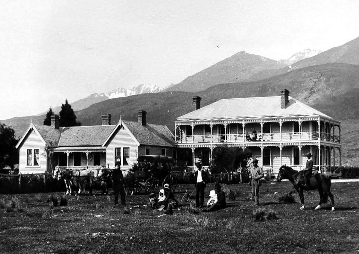 Photograph of a two-storey hotel with lace-trimmed verandahs beside a gabled cottage, a coach and horses, a saddled horse and a family on the tussock in front, mountains behind