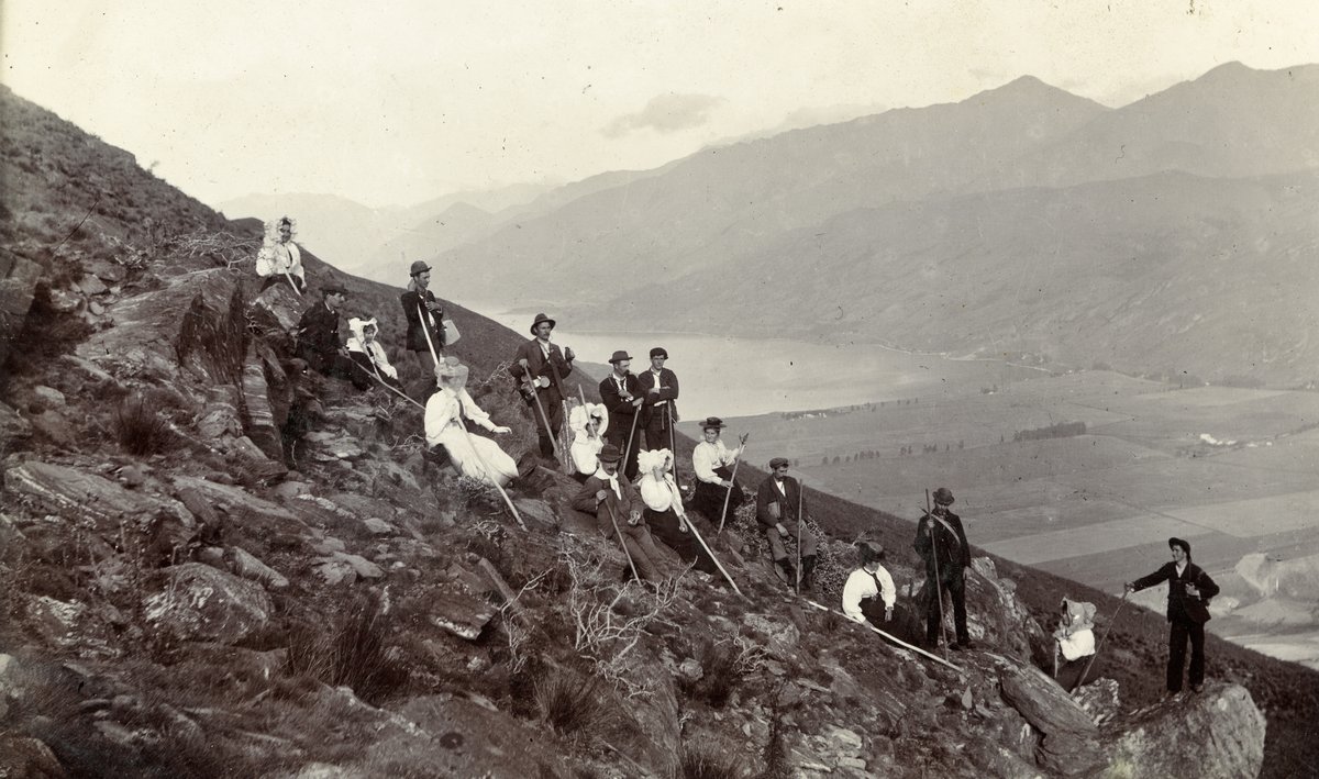 Photograph of about fifteen men and women posed on a steep rocky tussock slope with long walking poles, the men in suits and hats and the women in white full-length dresses and sunbonnets, the lake and the patchwork of Frankton Flats far below
